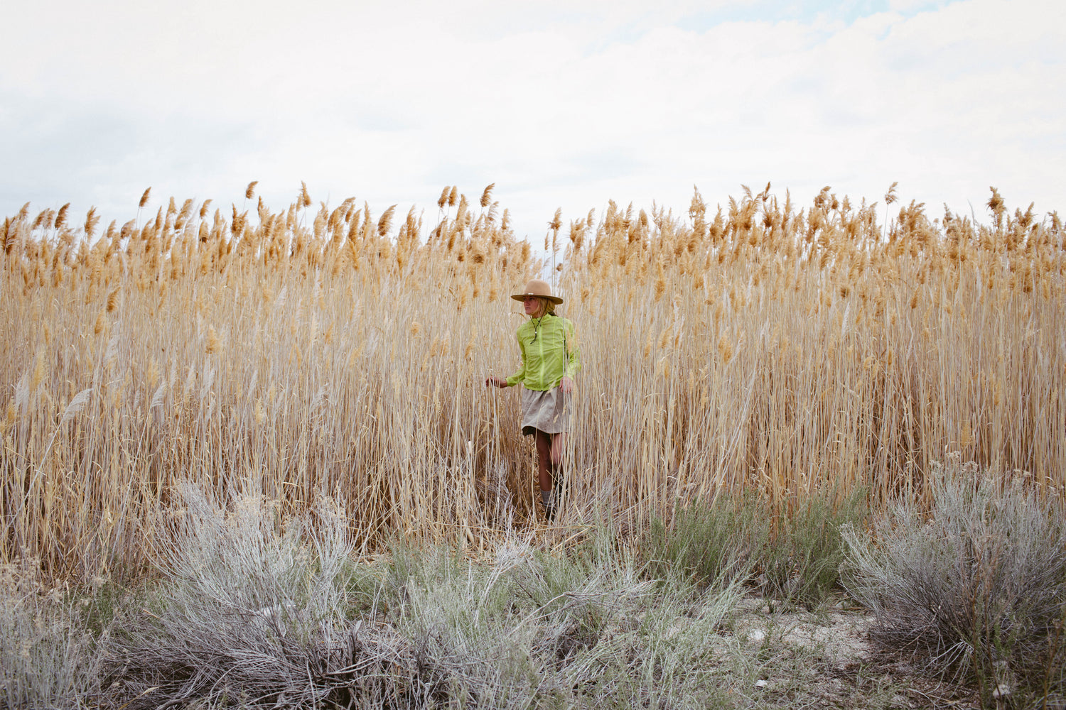 person exiting high a field of high grass, wearing a beige skirt, neon yellow windbreaker and felt hat