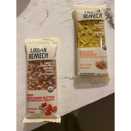 Two protein bars on a textured surface: Urban Remedy Cherry Cacao Peanut Butter Bar and Westerlind Golden Turmeric Bar, both labeled organic, plant-based, gluten-free, and non-gmo.