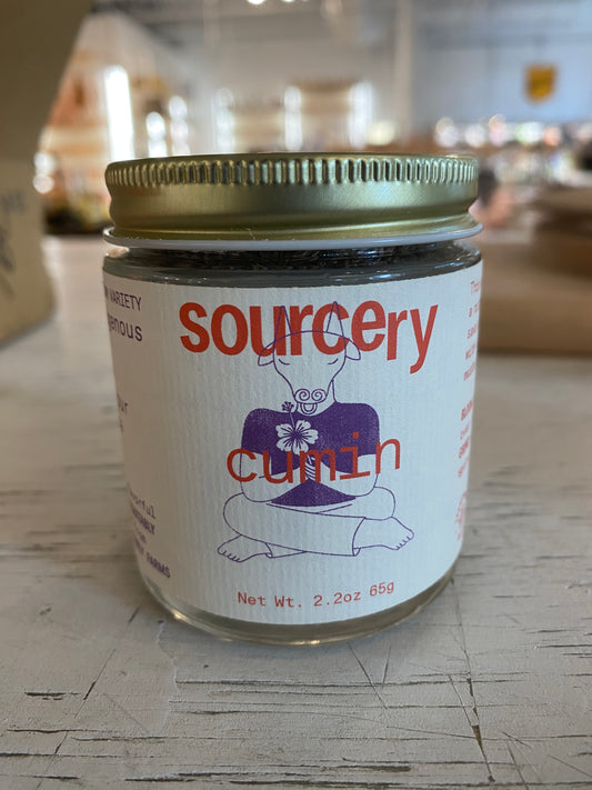A glass jar labeled "Sourcery Cumin" by Westerlind, with a gold lid and a figure in a flower shirt on the label, sits on a table. It has a net weight of 2.2 oz (65g).