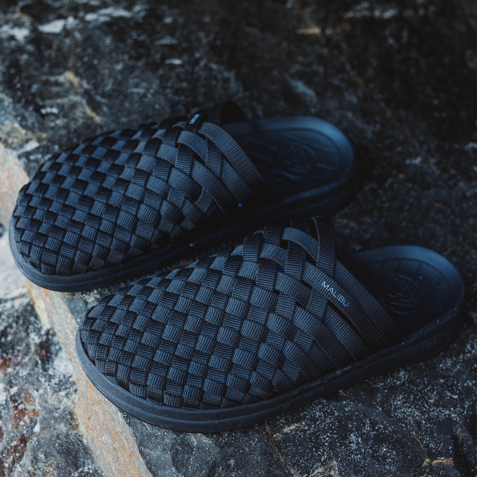 The Malibu Sandals W Colony: black hand-woven mule slip-ons with ergonomic footbed, displayed on a dark, textured stone surface.