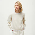 A person with blond hair wears the Inga Sweater by Aiayu, a smooth-knitted cream cashmere piece, paired with white pants. They stand against a plain light background, gaze turned to the side, hands in pockets.