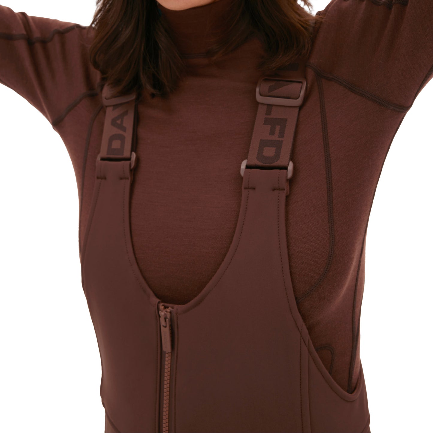 A person wearing a brown turtleneck is styled with the Halfdays Isabel Soft Shell Bib Pant, which features slim-fit design and adjustable straps. The pants have a front zipper, enhancing their sleek and stylish appearance.