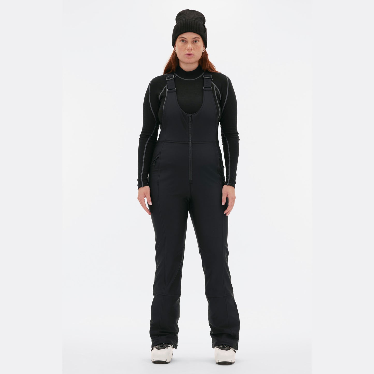 A person wearing a black beanie, black thermal clothing, and slim-fit Isabel Soft Shell Bib Pant by Halfdays stands against a plain white background.
