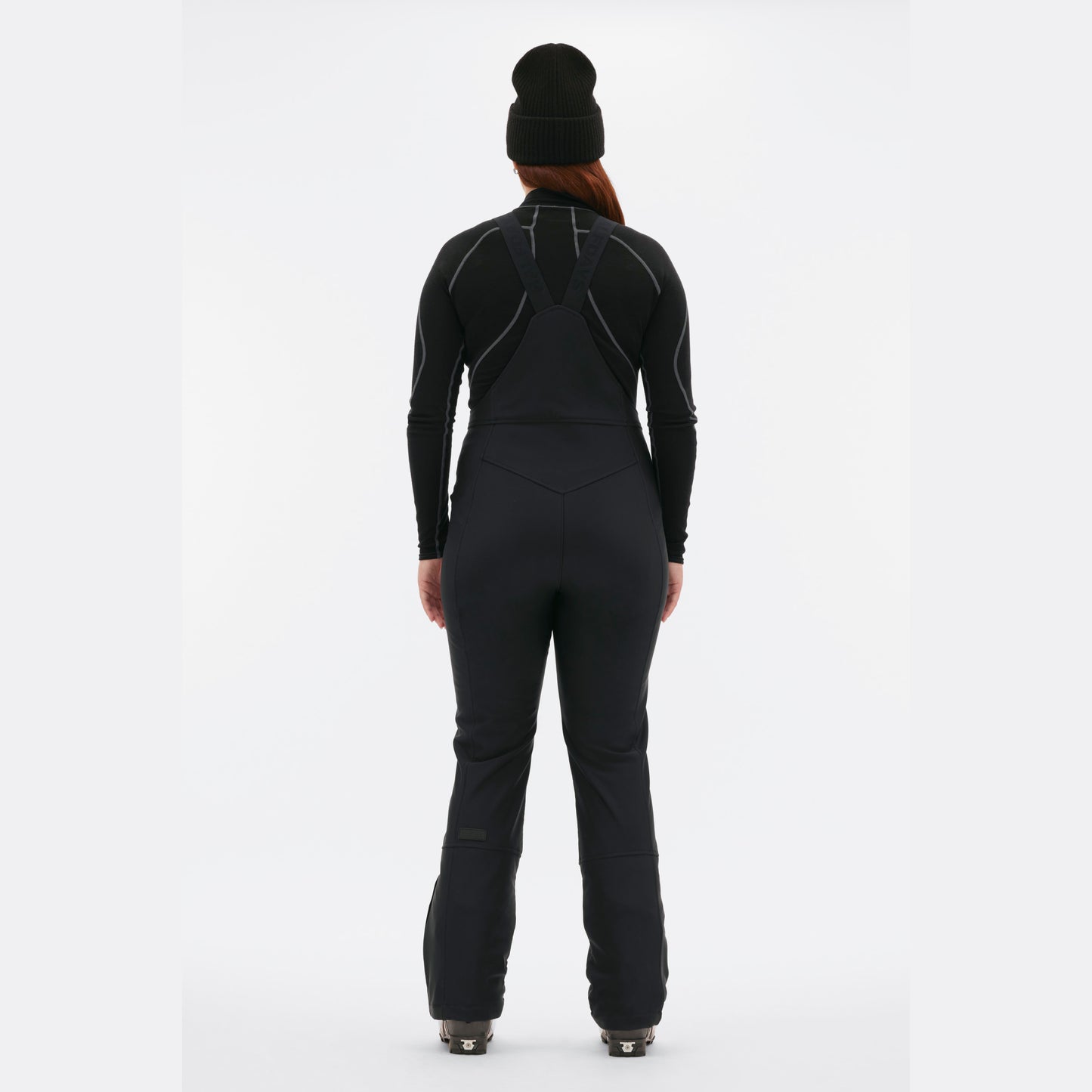 A person wearing Halfdays' Isabel Soft Shell Bib Pant in black slim-fit winter style and a beanie stands with their back to the camera against a white background, showcasing the sleek design of the gear.