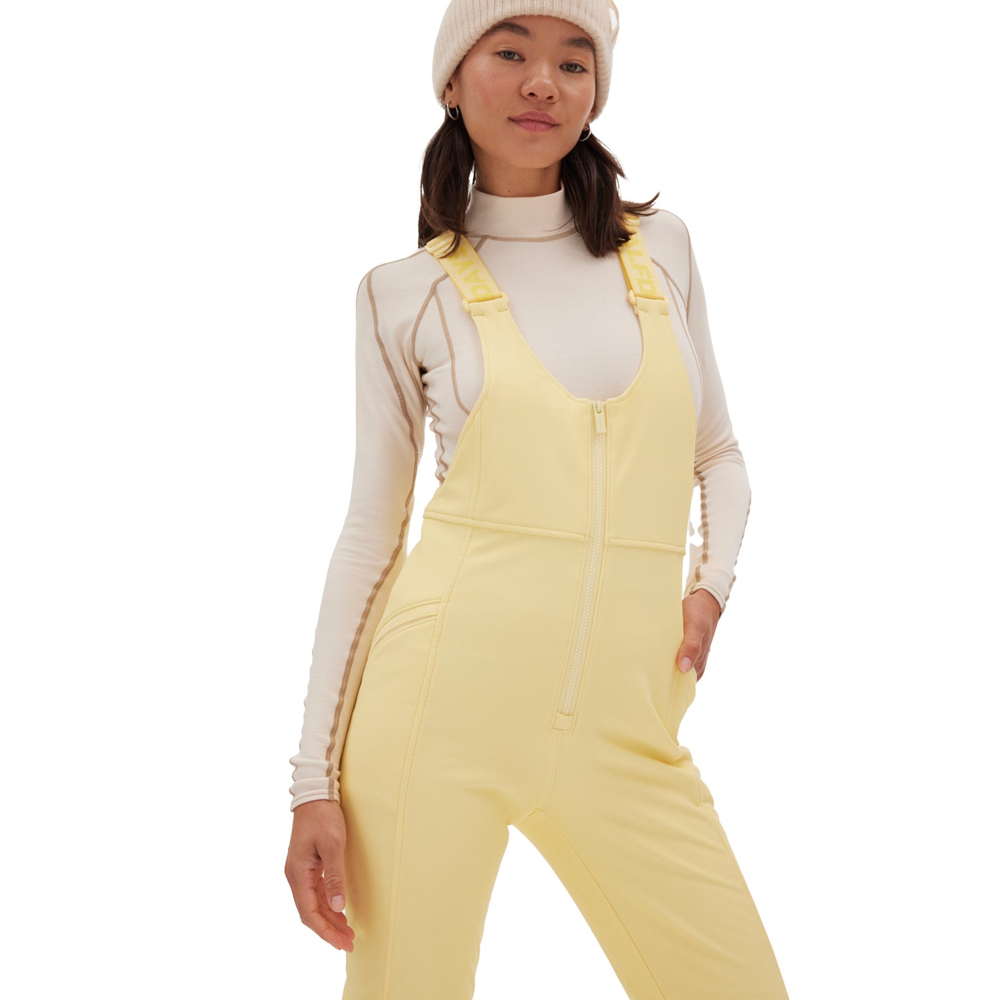 A person wearing a white turtleneck, the Isabel Soft Shell Bib Pant in a slim-fit yellow design by Halfdays, and a white beanie poses against a plain background.