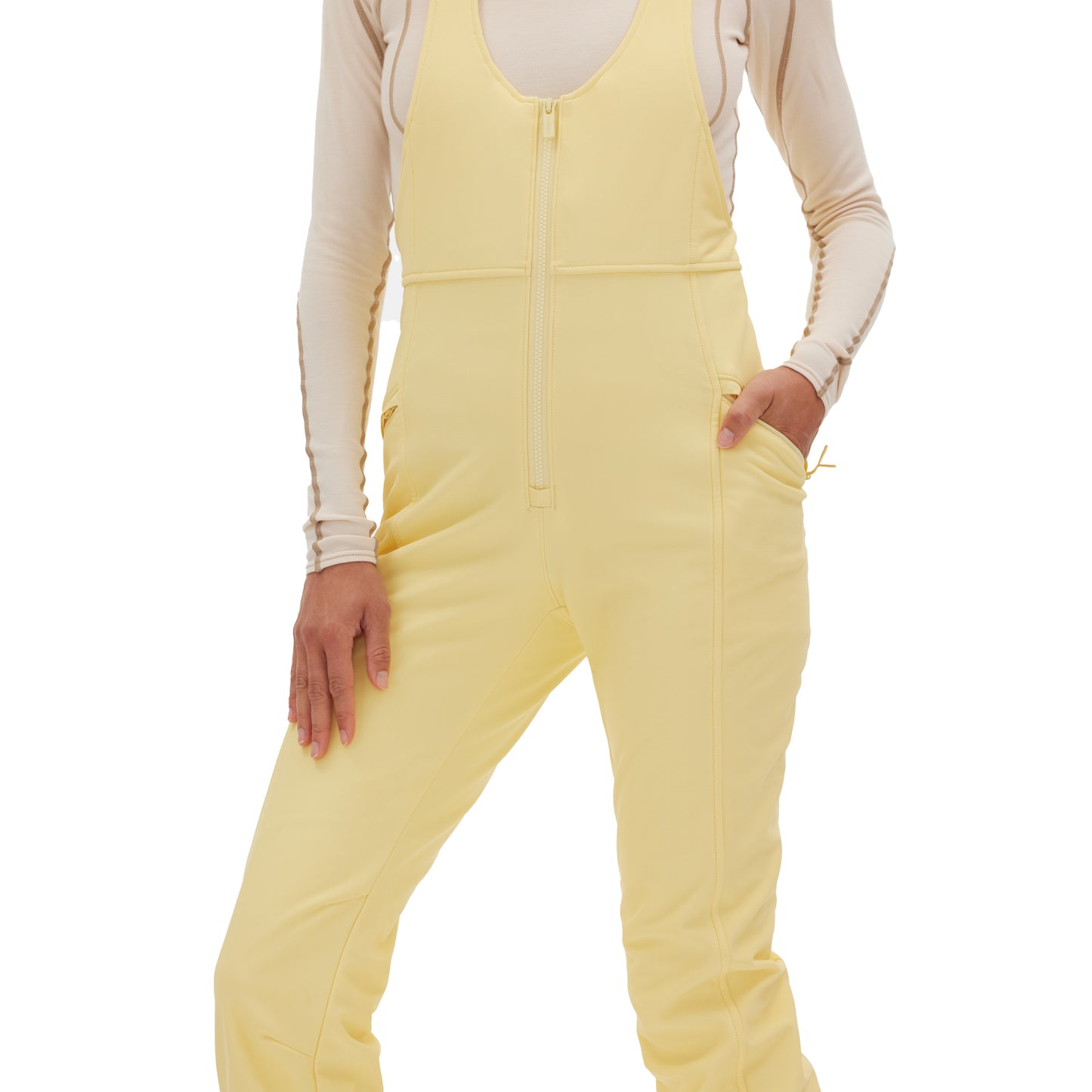 A person wearing the Isabel Soft Shell Bib Pant from Halfdays, featuring a light yellow color with a zip-front and pockets in a slim-fit design, layered over a long-sleeved beige top.