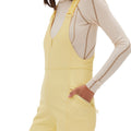 A person wearing the Isabel Soft Shell Bib Pant by Halfdays, a yellow slim-fit design similar to a soft shell style, over a beige long-sleeve top. The jumpsuit includes a front zipper and pockets.