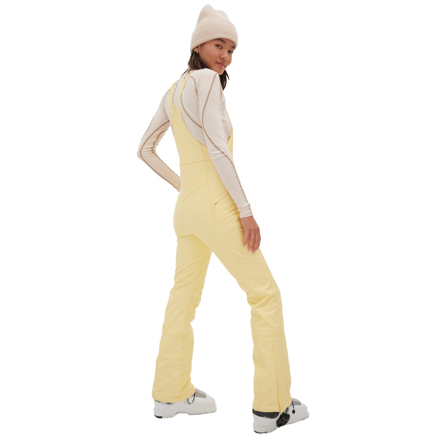 A person confidently looks over their shoulder, one hand on their hip, wearing a cream beanie and long-sleeve top paired with the yellow Isabel Soft Shell Bib Pant from Halfdays.