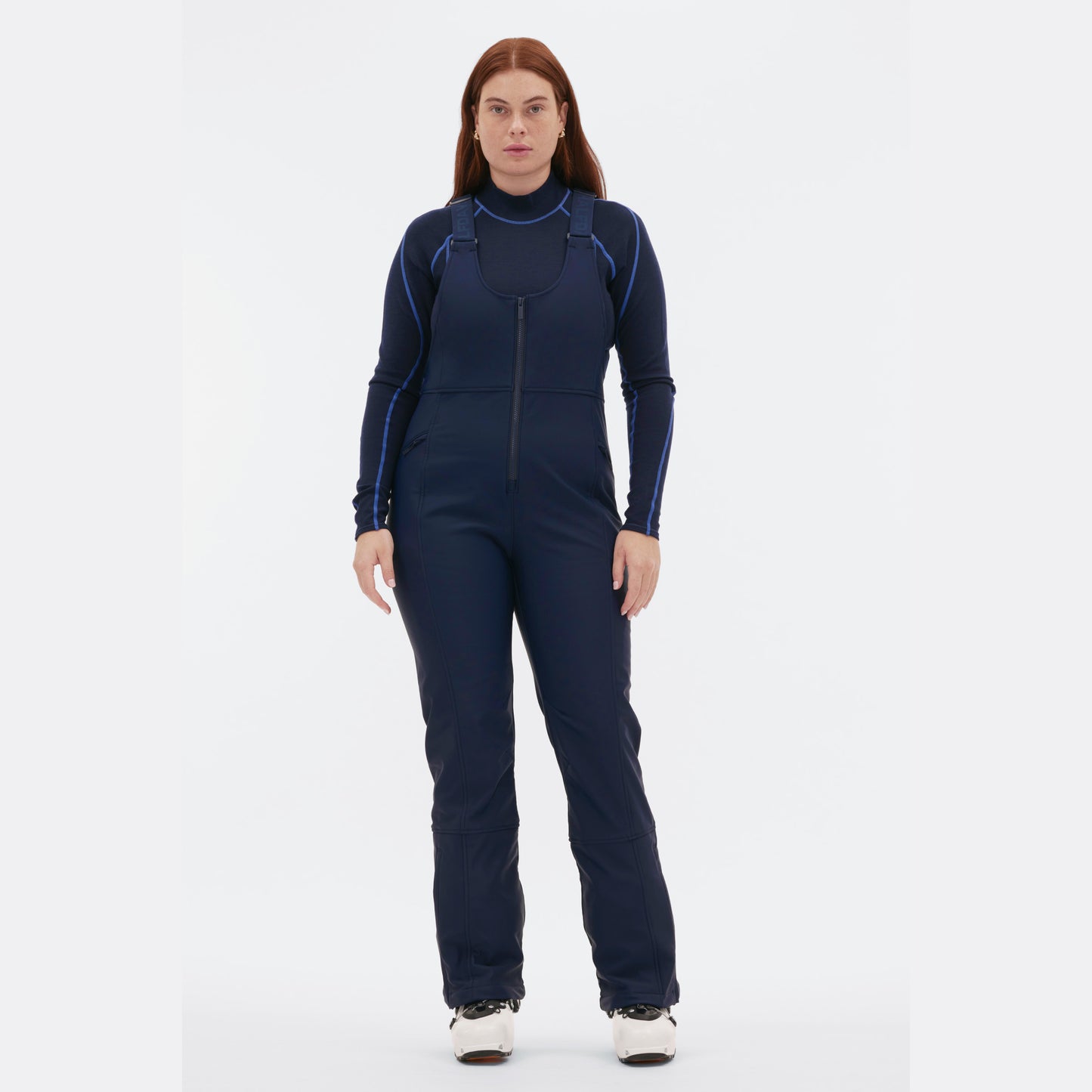 Wearing the Isabel Soft Shell Bib Pant by Halfdays, a person in a navy-blue ski suit and white boots with a slim-fit design stands against a plain white backdrop.