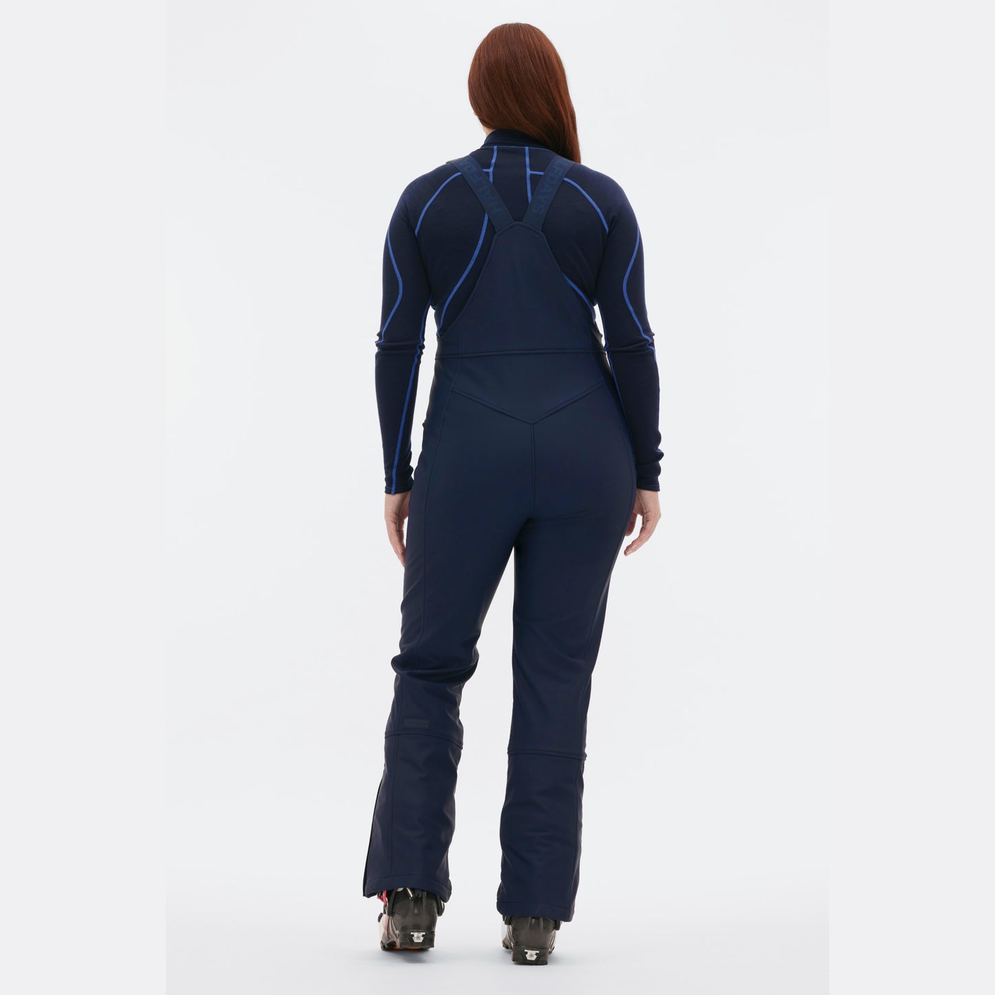 A person stands with their back to the camera in a sleek dark blue Halfdays Isabel Soft Shell Bib Pant, highlighting its slim-fit design.
