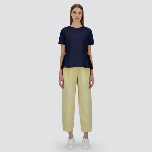 A woman stands facing forward against a plain light gray background, wearing Herno's Mix Taffeta Ruffle Back T-Shirt in navy blue, paired with light yellow wide-leg pants and white sneakers.