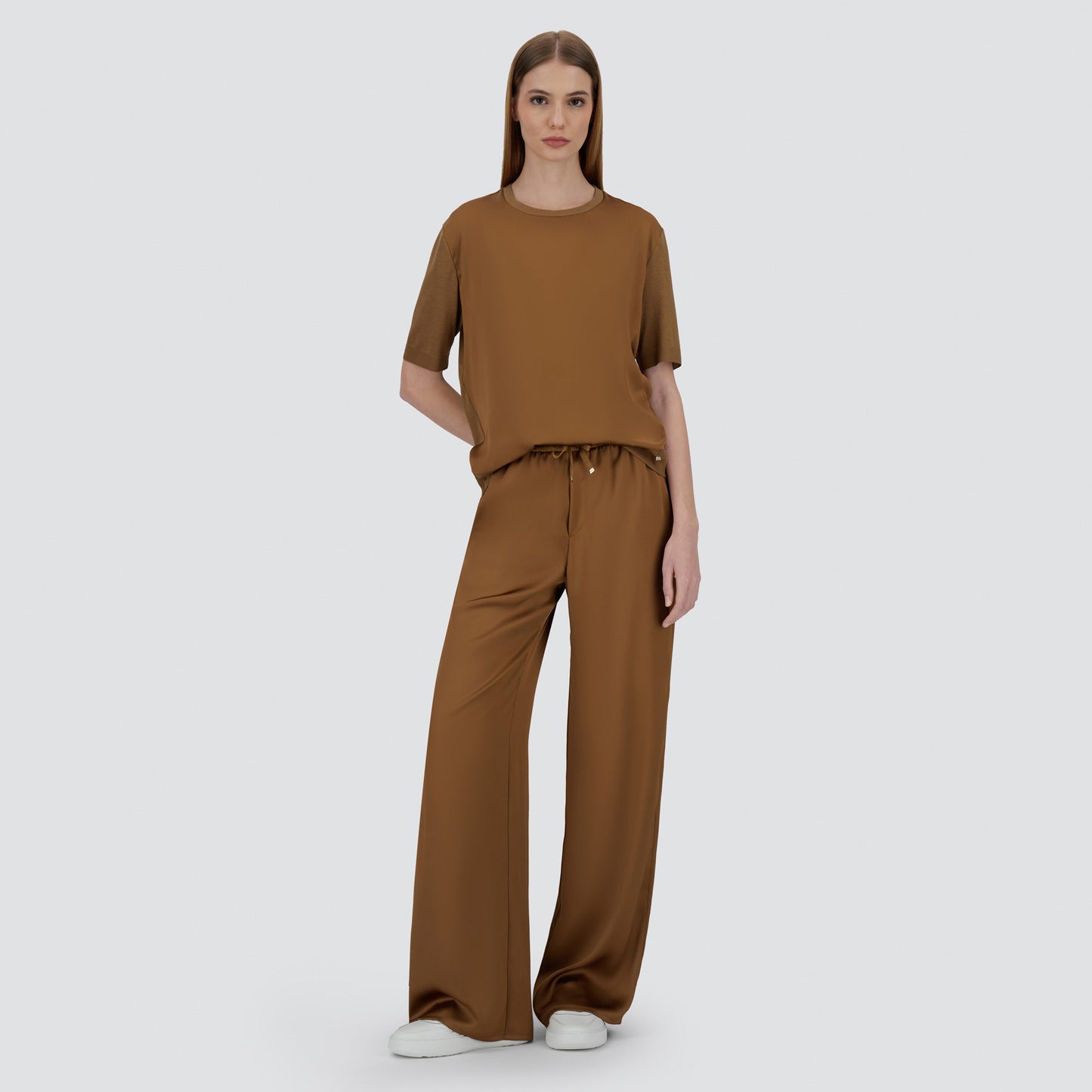A woman stands against a plain background wearing the Herno Satin Front T-Shirt in brown and matching wide-leg pants, paired with white sneakers.