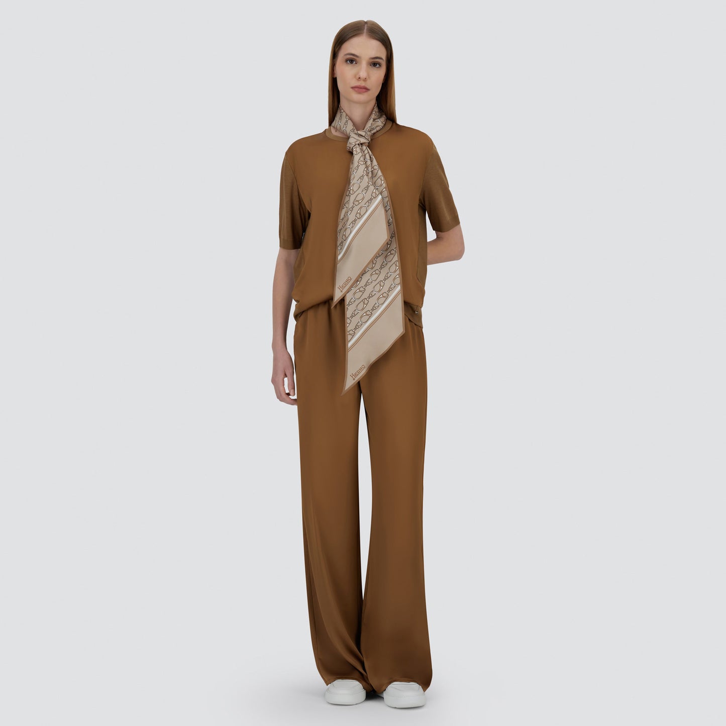 A woman faces forward against a plain light background, wearing the Herno Satin Front T-Shirt in brown, wide-leg pants to match, a beige patterned scarf, and white shoes.
