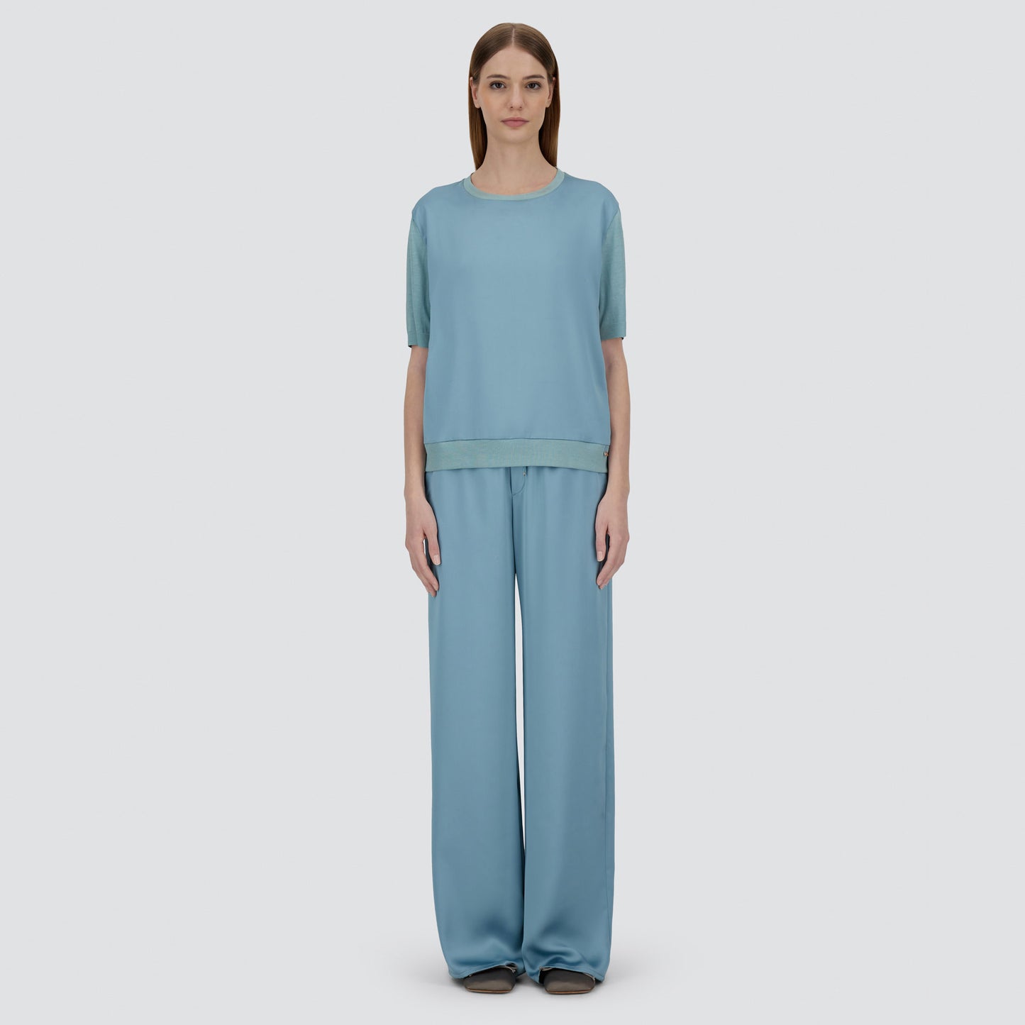 A woman faces forward against a plain light background, wearing Herno’s Satin Front T-Shirt in light blue with matching wide-leg pants.