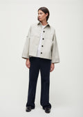 A person is wearing a Jules Jacket by Aiayu, made from light-colored, weather-resistant Ventile fabric, paired with dark pants and black shoes against a plain background.