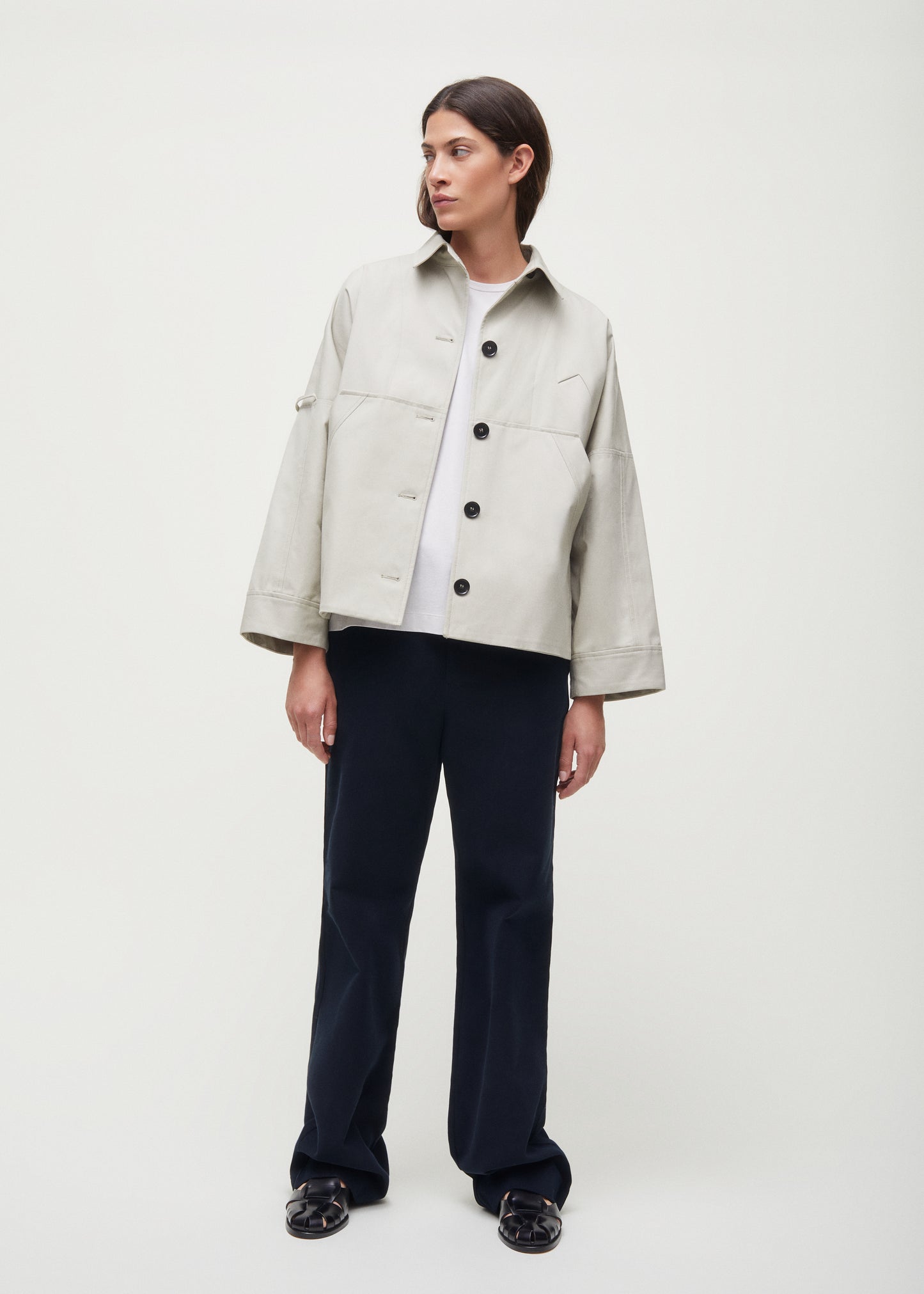 A person is wearing a Jules Jacket by Aiayu, made from light-colored, weather-resistant Ventile fabric, paired with dark pants and black shoes against a plain background.