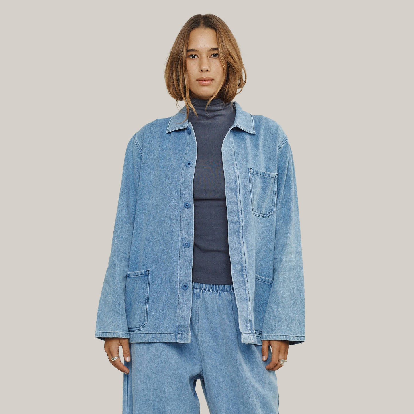 A person in the Jungmaven Olympic Jacket in Light Denim styled with matching pants and a dark turtleneck stands against a plain light background.