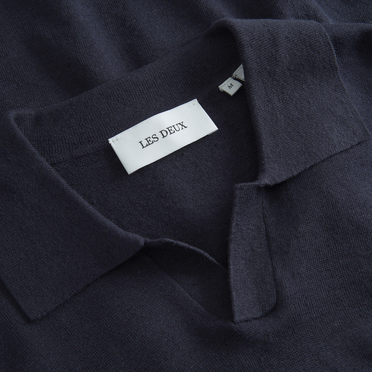 A close-up of the Les Deux Knit Polo in dark navy, showcasing vintage style with a collar and "Les Deux" branding plus an "M" size tag near the neckline.