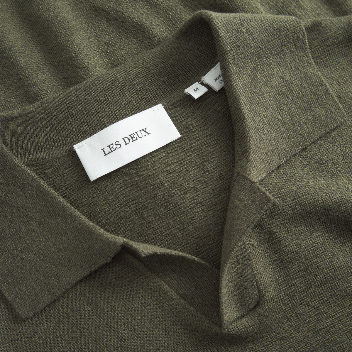 Close-up of a Knit Polo in olive green from Les Deux, showcasing its vintage-inspired style with the brand’s label and size M tags visible.