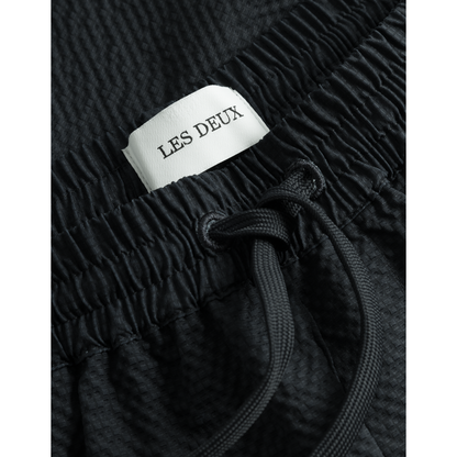 Close-up of a black textured Stan Seersucker Swim Shorts 2.0 made of quick-dry seersucker material, with a white label reading "Les Deux" and a black elasticated drawstring waist.