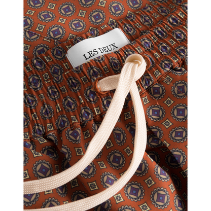 Close-up of a garment with orange fabric and intricate blue patterns, featuring a white 'Stan AOP Swim Shorts 2.0, Terrocotta/Pineapple' label and a beige drawstring waist by Les Deux.