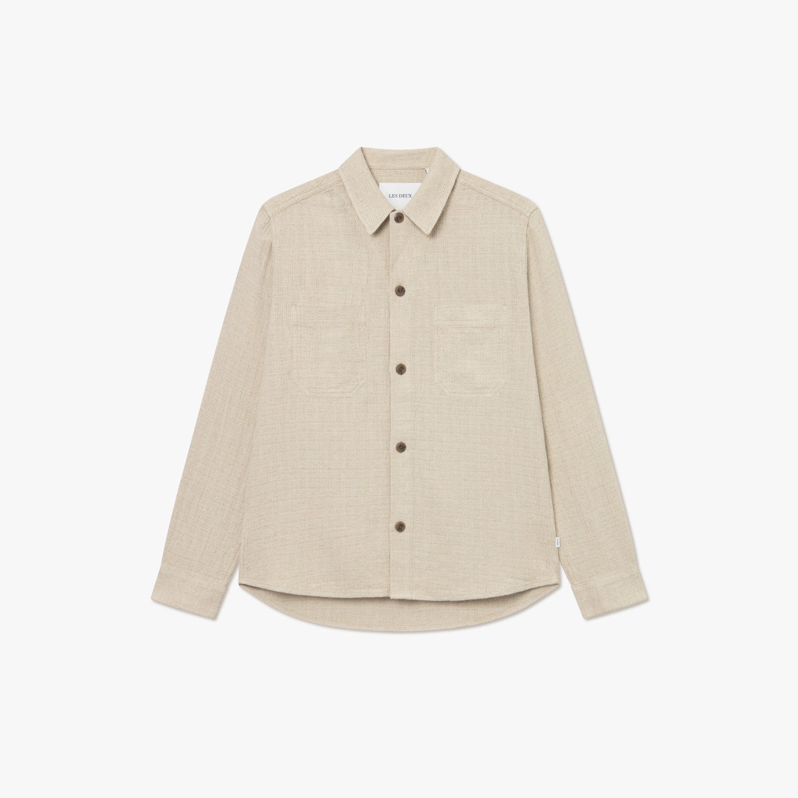 The Les Deux Hamilton Overshirt is a beige long-sleeve button-up made from a breathable linen blend, featuring two chest pockets, a spread collar, and brown buttons. Shown on a white background.