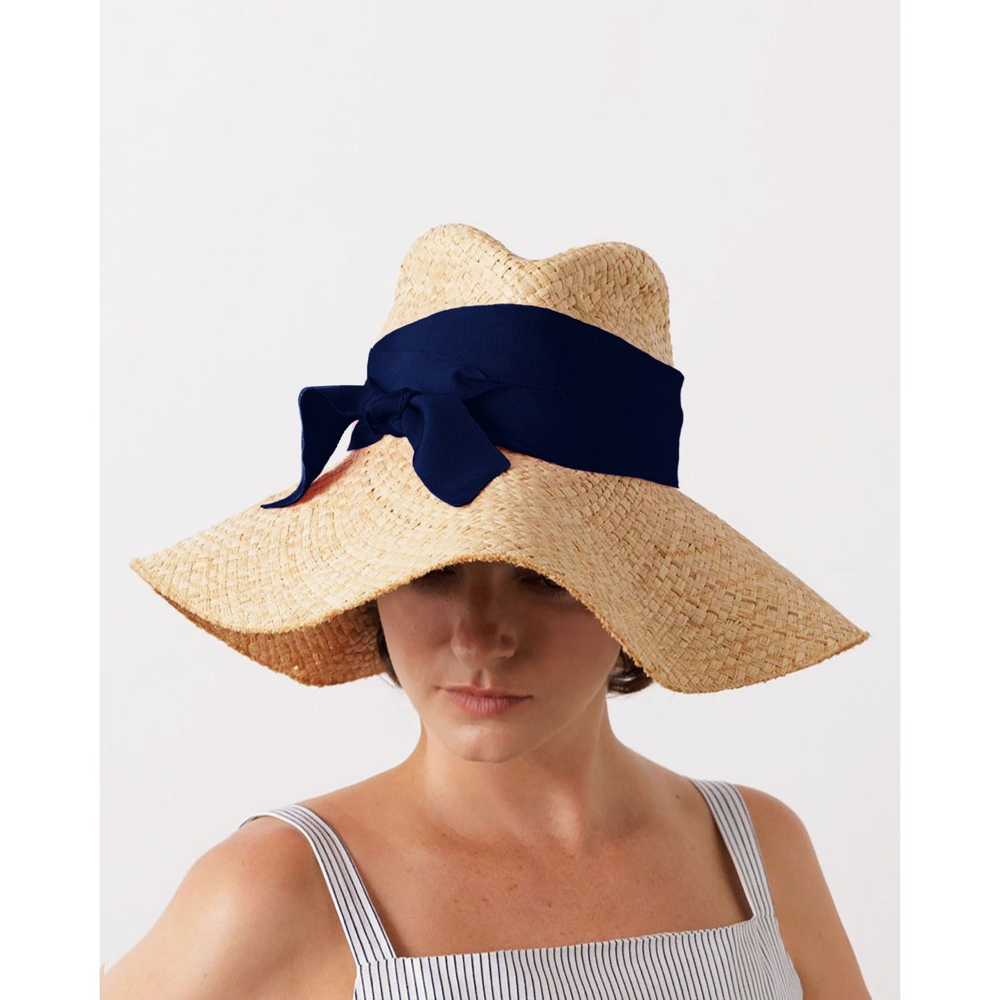 A person wears the First Aid, Nude w/ Navy by Lola Hats, adorned with a dark blue ribbon and bow, perfectly paired with a striped top. This summer style image is set against a plain white background.