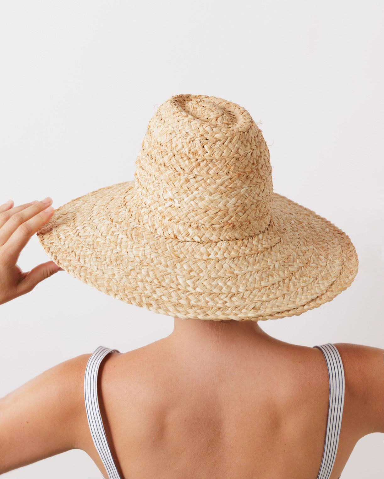 A person seen from behind wearing a wide-brimmed raffia hat (Fiscolo, Natural) by Lola Hats, with a hint of striped clothing visible on the shoulders.