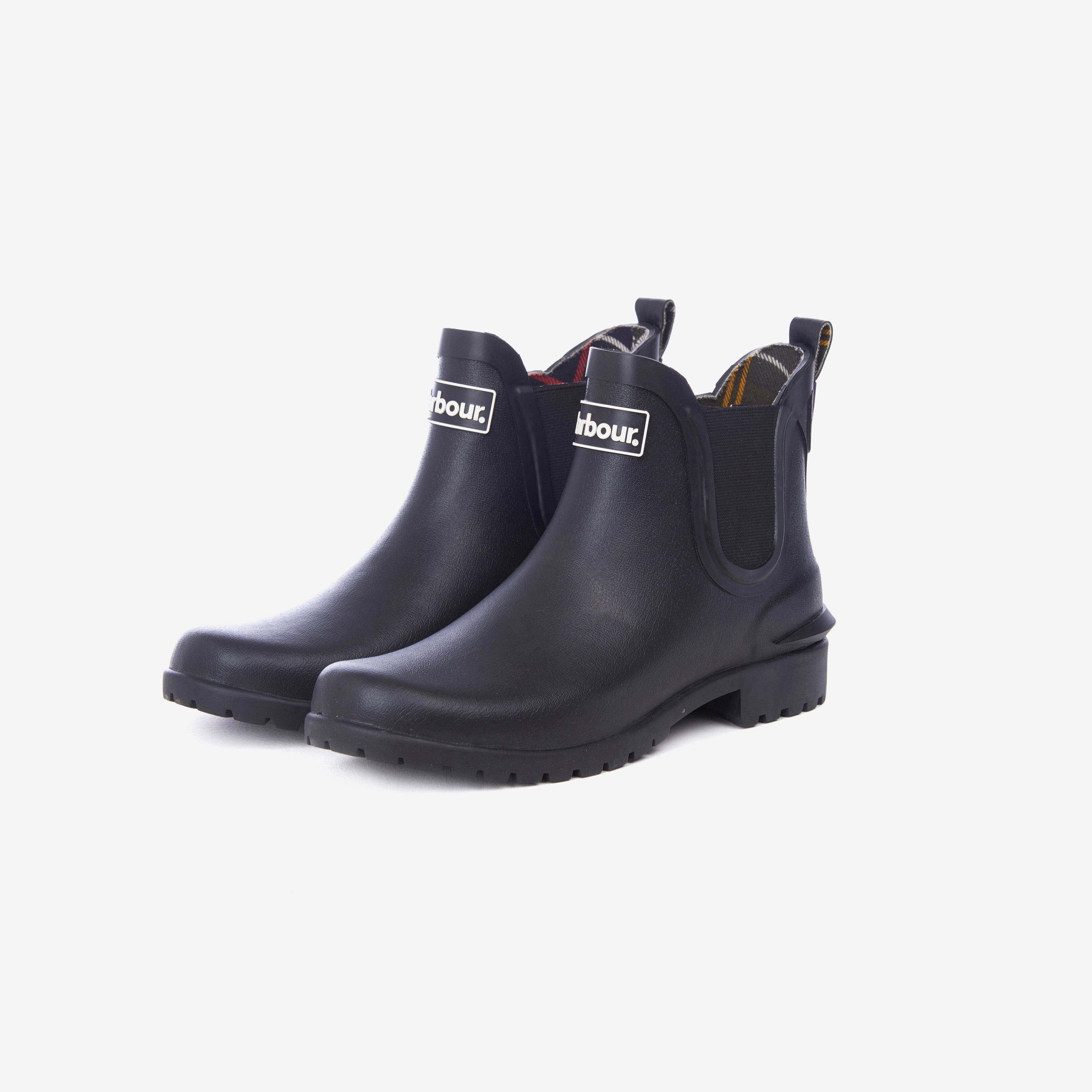 The Barbour Wilton Chelsea Welly is a pair of black ankle-length boots with pull tabs, a front Barbour logo, and tartan lining, shown on a white background.