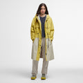 A person models the Barbour Evie Showerproof Parka in yellow with an adjustable hood over a black shirt and beige pants, paired with matching yellow shoes, against a plain white backdrop.