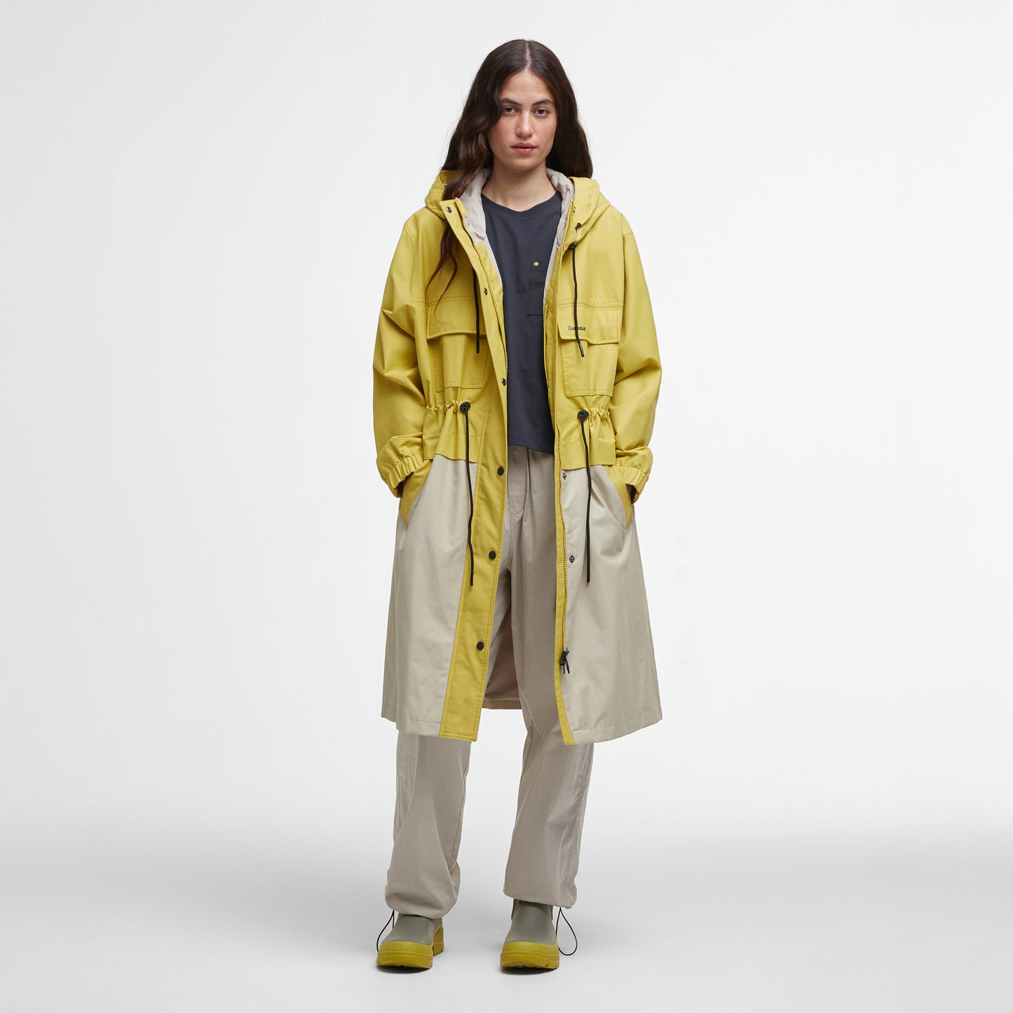 A person models the Barbour Evie Showerproof Parka in yellow with an adjustable hood over a black shirt and beige pants, paired with matching yellow shoes, against a plain white backdrop.
