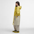 A person wearing the Barbour Evie Showerproof Parka with an adjustable hood stands in profile, paired with beige pants, against a plain background.