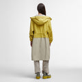 A person, clad in a Barbour Evie Showerproof Parka in yellow and beige with an adjustable hood, pairs it with matching pants and boots while standing against a plain background, facing away.