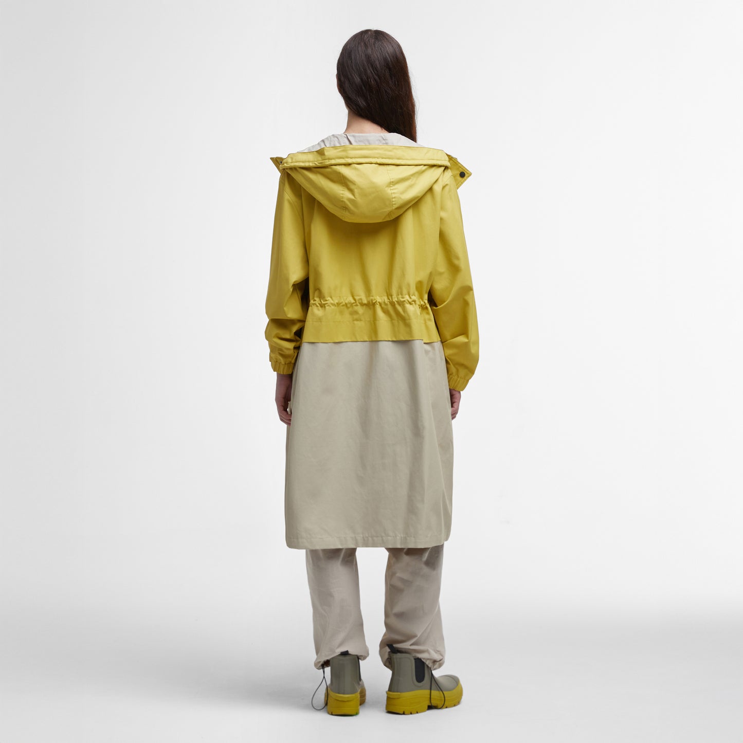 A person, clad in a Barbour Evie Showerproof Parka in yellow and beige with an adjustable hood, pairs it with matching pants and boots while standing against a plain background, facing away.