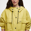 A person in a yellow Barbour Evie Showerproof Parka with an adjustable hood and black drawstrings is standing against a plain white background.