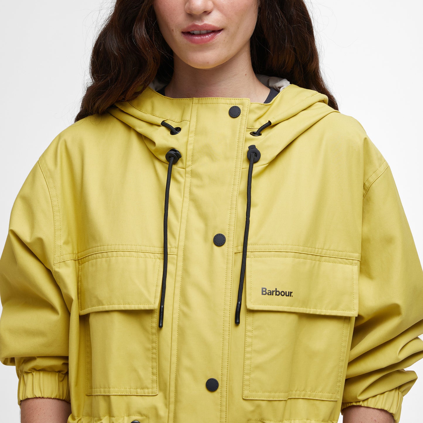 A person in a yellow Barbour Evie Showerproof Parka with an adjustable hood and black drawstrings is standing against a plain white background.