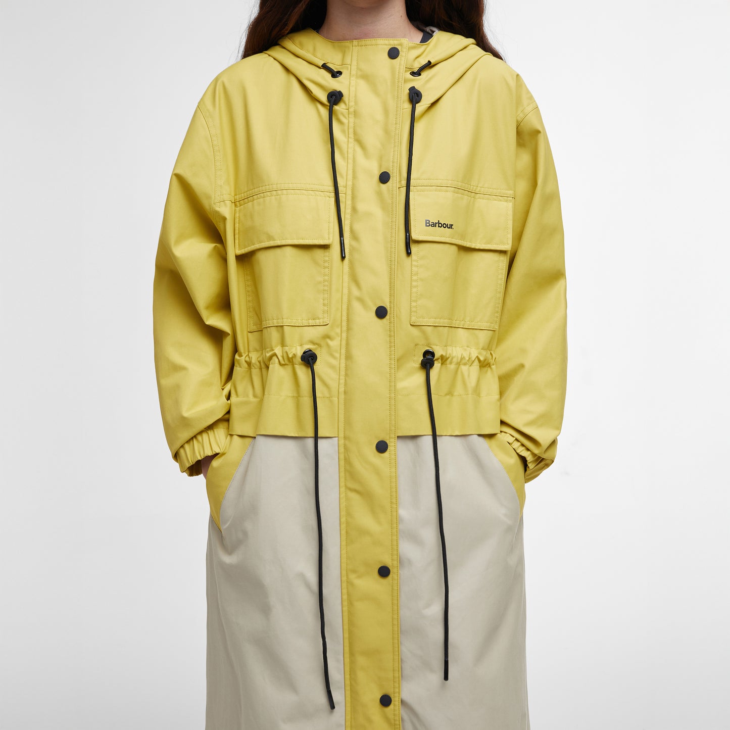 The person is wearing the Barbour Evie Showerproof Parka, a yellow and beige hooded jacket with black drawstrings and buttons, providing excellent weather protection.
