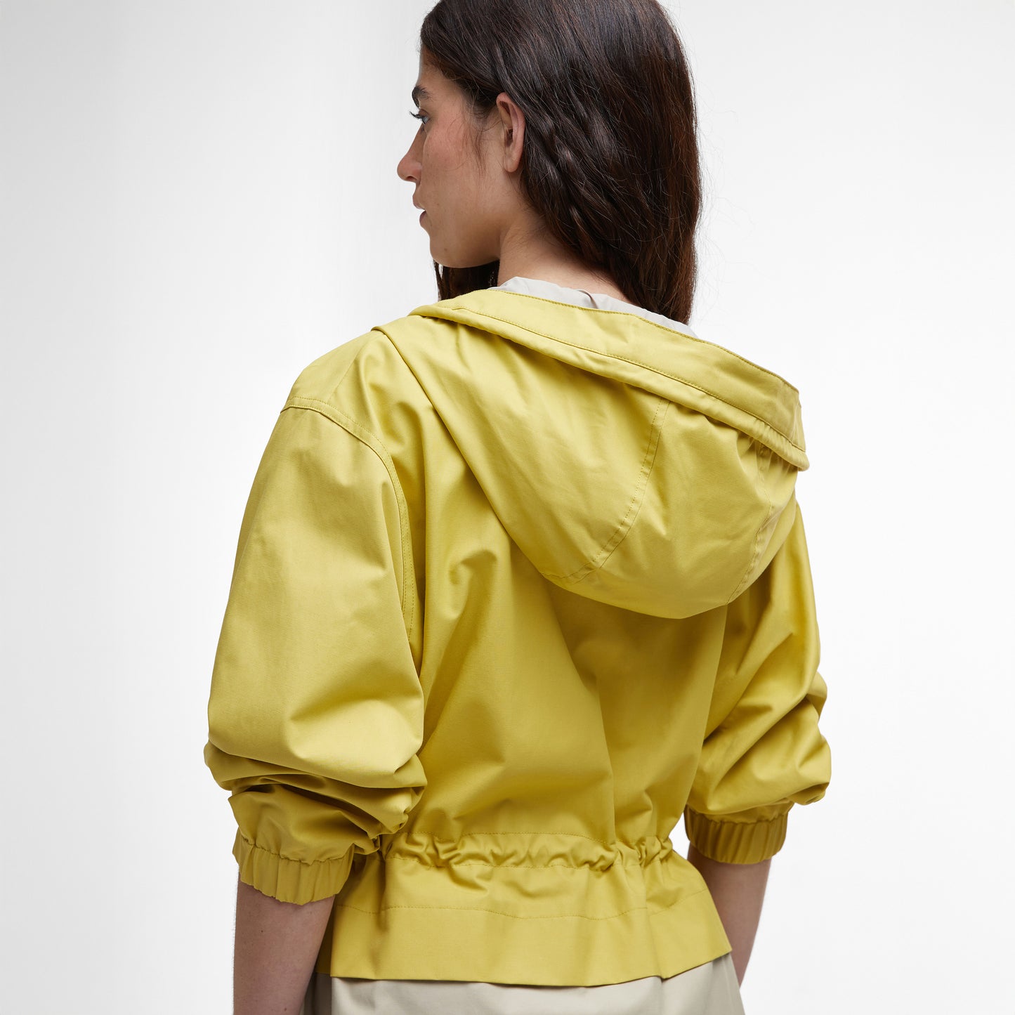 Facing away, a person wears the Barbour Evie Showerproof Parka in yellow. The photo focuses on their upper body against a light gray background, emphasizing the parka's adjustable hood and weather protection features.