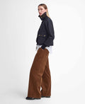 A person stands sideways, wearing a Barbour W Cropped Beadnell Waxed Jacket over a white shirt, wide-leg brown corduroy pants, and black shoes, against a plain white background.