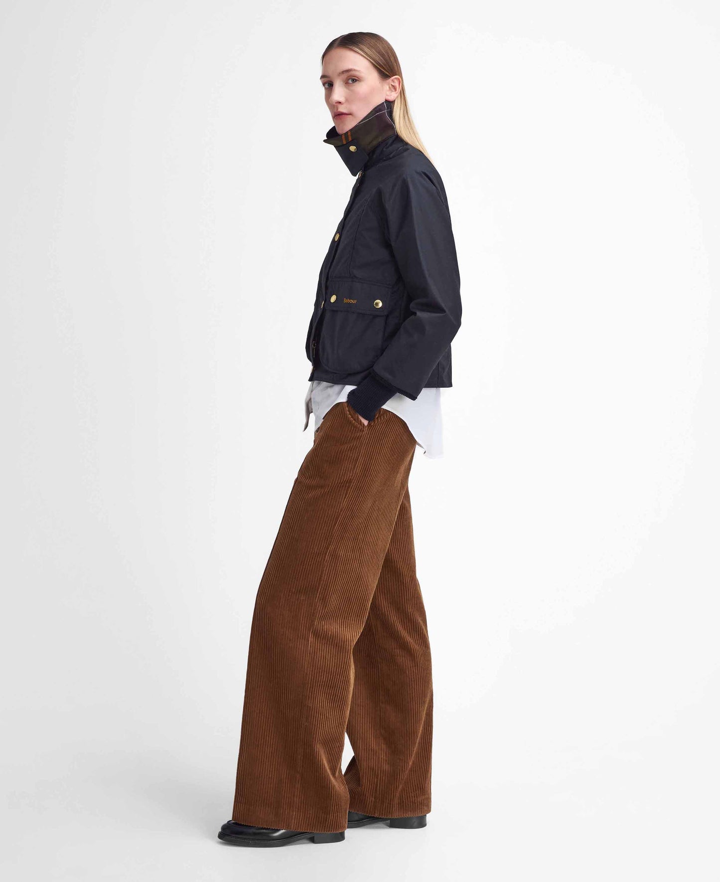 A person stands sideways, wearing a Barbour W Cropped Beadnell Waxed Jacket over a white shirt, wide-leg brown corduroy pants, and black shoes, against a plain white background.