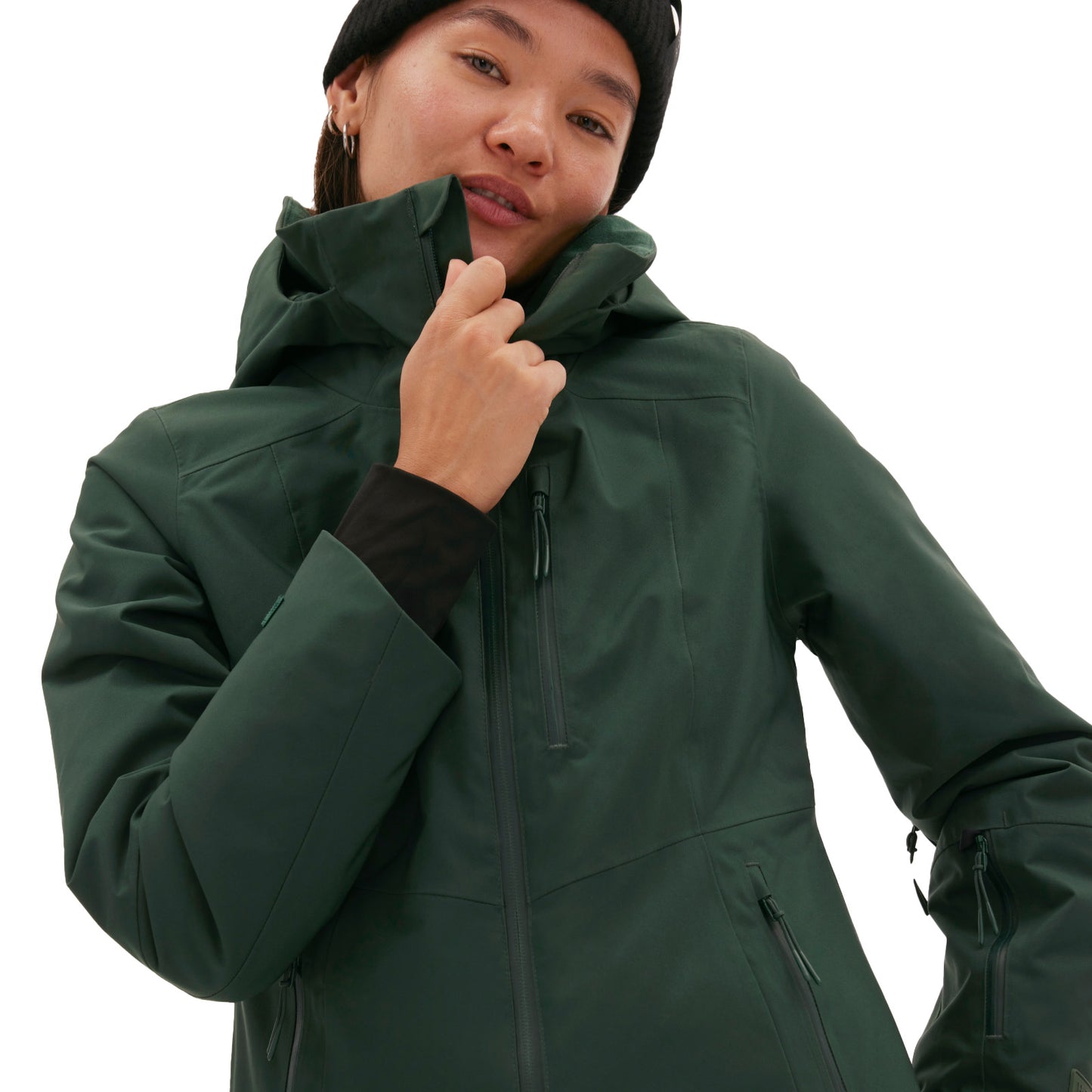 A woman wearing the Halfdays Lawrence Jacket in dark green and a black beanie holds the collar up against a plain background.