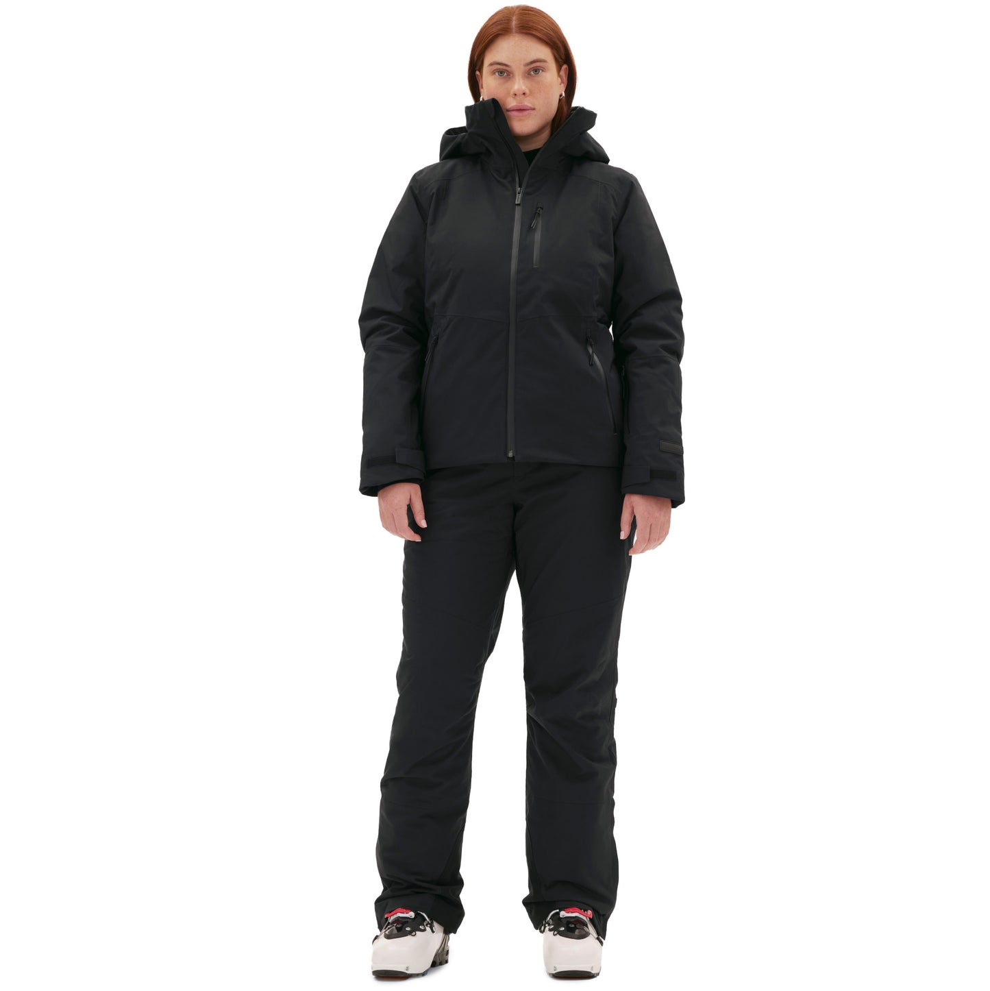 Against a white backdrop, a woman stands confidently in her ski boots, clad in the Lawrence Jacket by Halfdays and black winter pants.