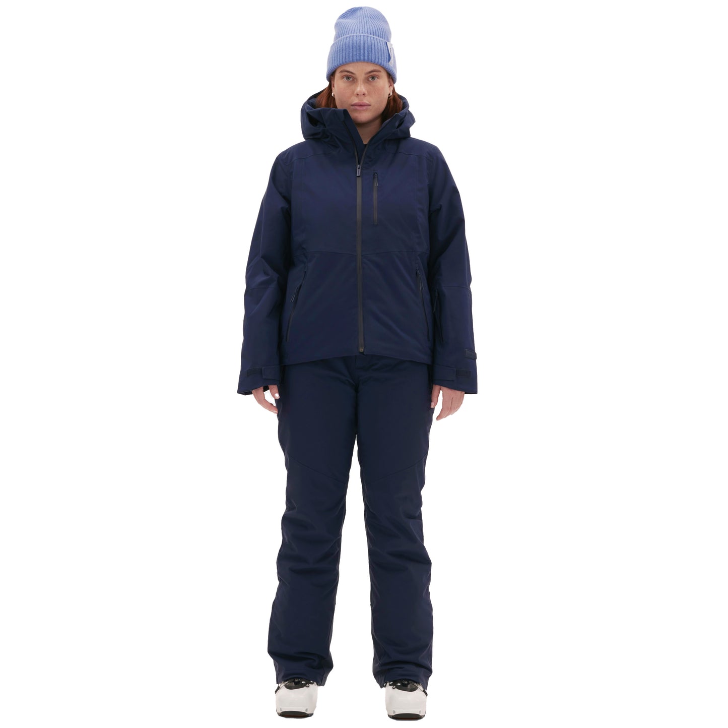 A confident woman poses against a plain white background, ready for the slopes in a navy Lawrence Jacket by Halfdays with matching pants, a light blue beanie, and white snow boots.