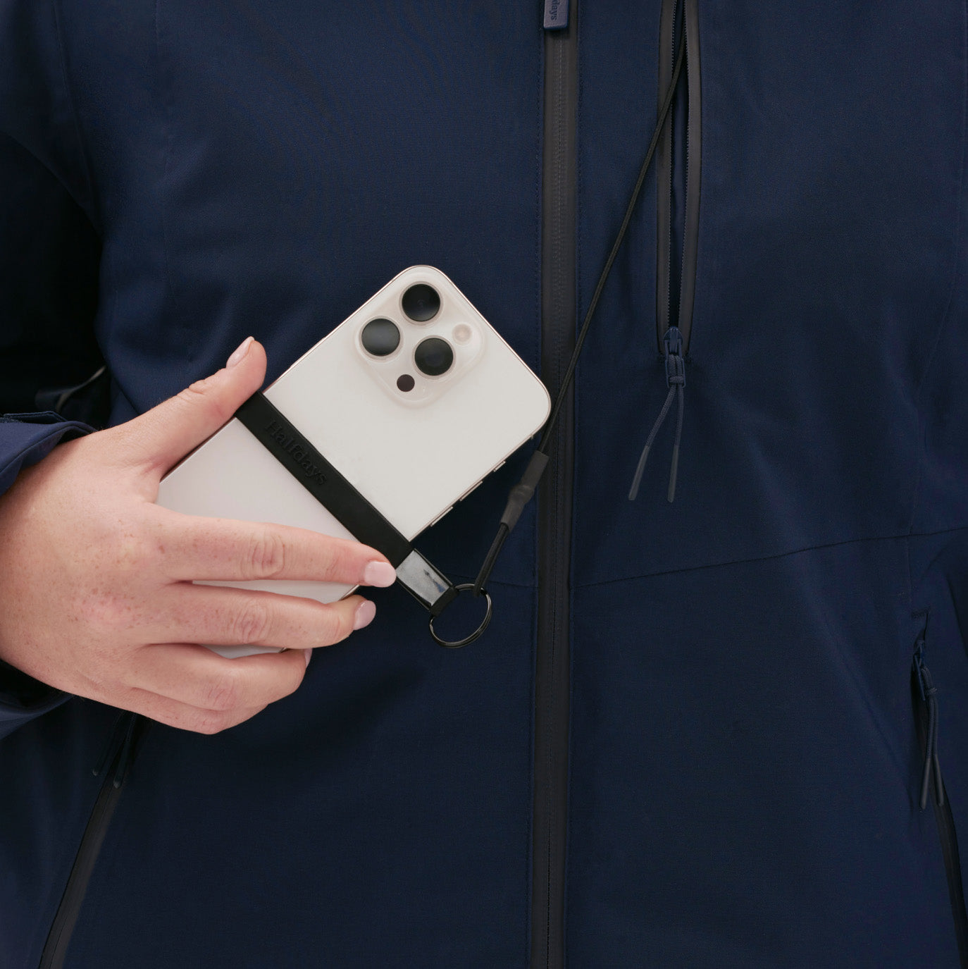 A woman holds a smartphone with a black strap, wearing the Lawrence Jacket by Halfdays, in dark blue.