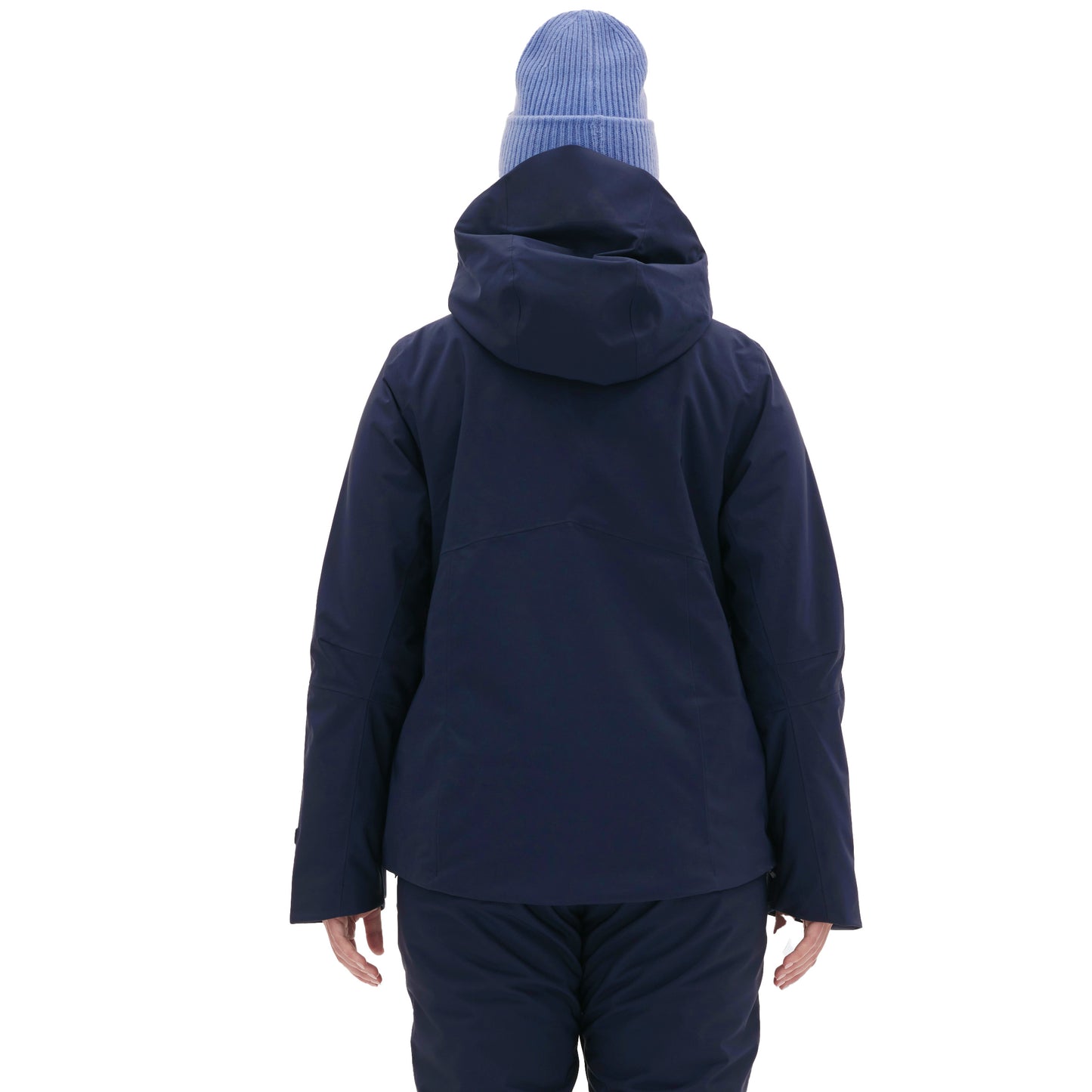 A woman in the Lawrence Jacket by Halfdays and a blue beanie stands with her back to the camera against a pristine white backdrop, conveying calm readiness for the slopes.