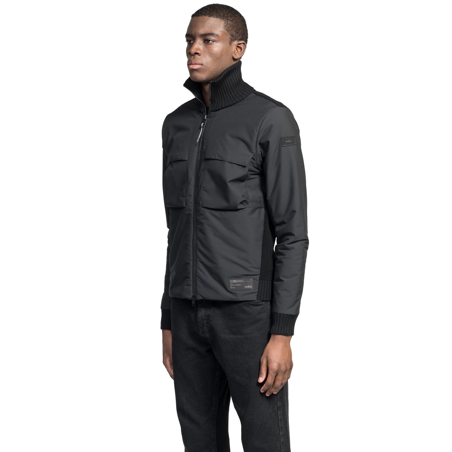 A person in a fitted black Layton Men's Tactical Hybrid Sweater by Nobis with Primaloft Gold Insulation and dark jeans stands against a white background, looking to the side.