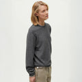 A person with short blonde hair is wearing a sleek, dark gray Aiayu Leonardo Cashmere Sweater and beige pants, standing against a plain light background.
