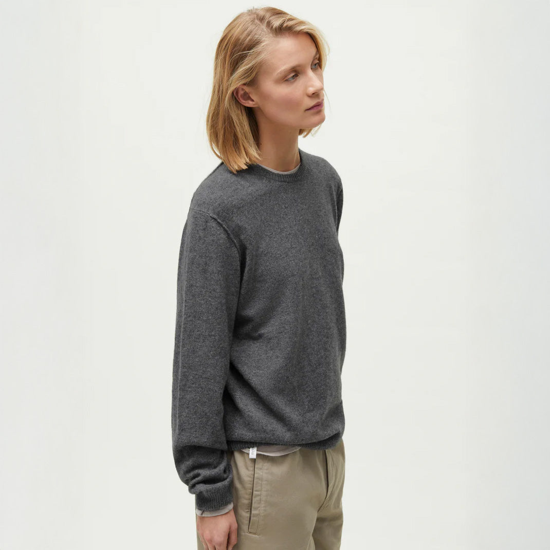 A person with short blonde hair is wearing a sleek, dark gray Aiayu Leonardo Cashmere Sweater and beige pants, standing against a plain light background.