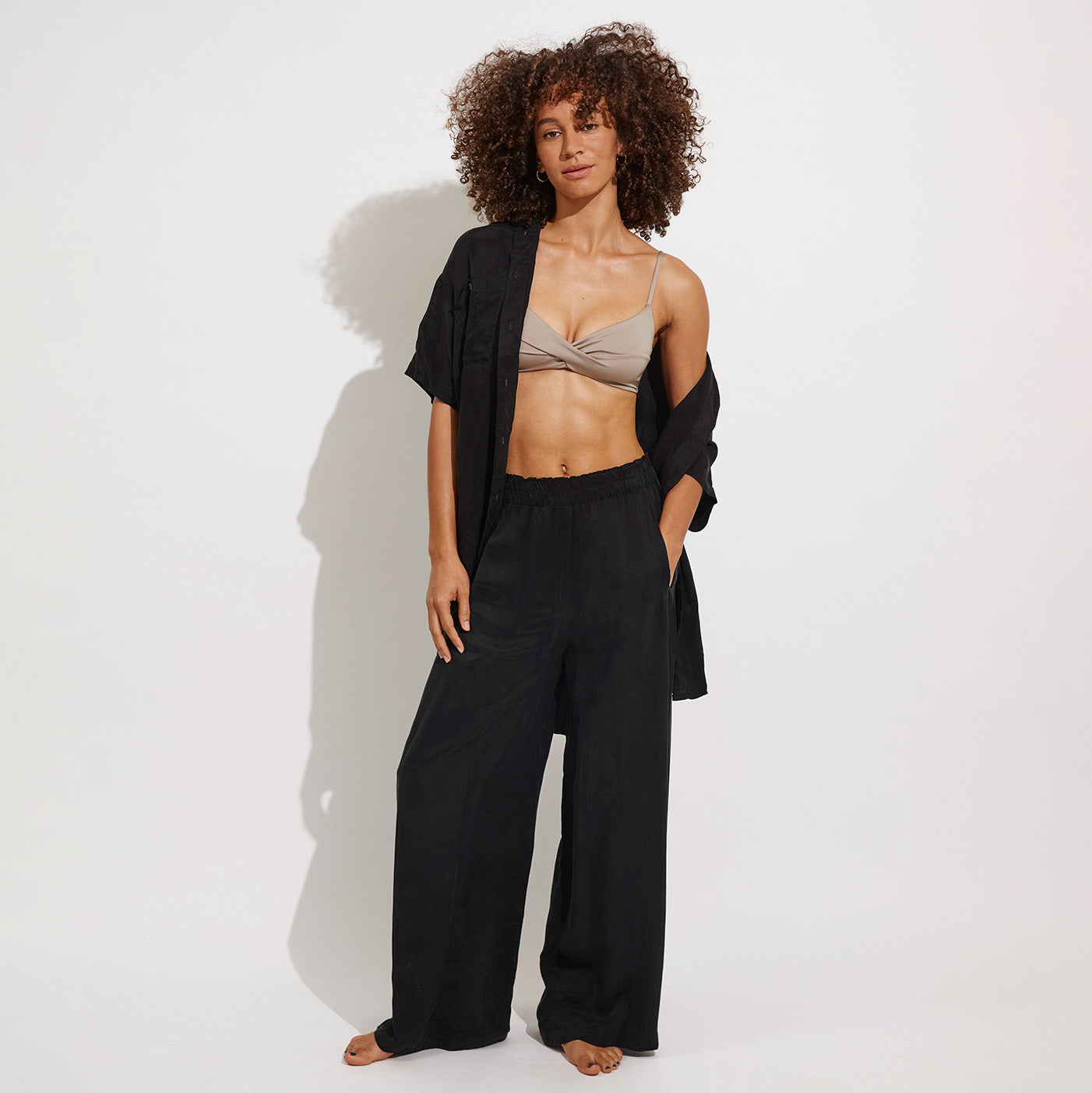 A woman stands barefoot against a white background, wearing a taupe bralette, black wide-leg pants, and the Left On Friday Day Trip Button Down—an oversized black shirt with a button-down front.