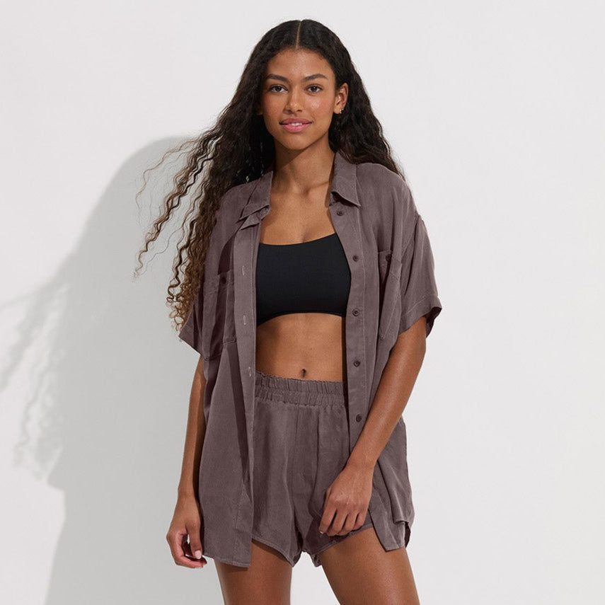 A woman with long curly hair wears the Left On Friday Day Trip Button Down in brown, matching shorts, and a black crop top, standing against a plain light background.